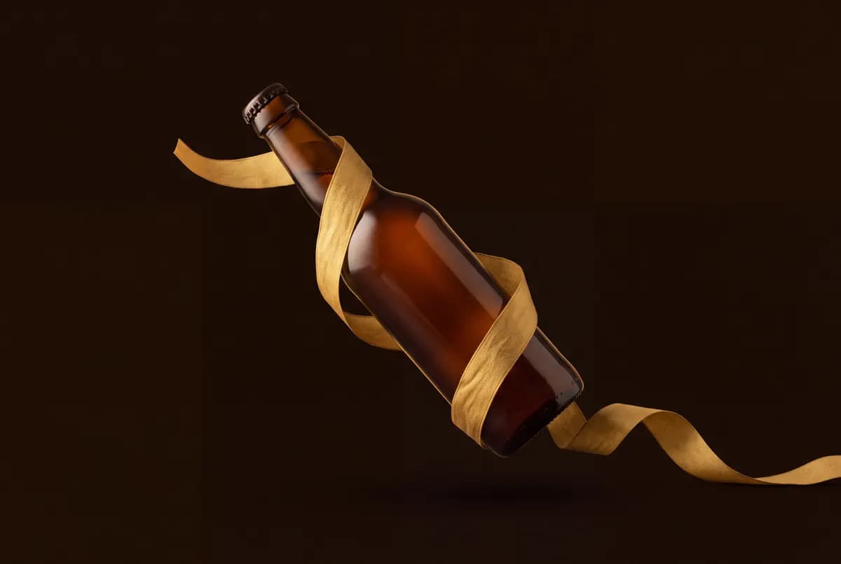 Bouteille de bière brune couchée en diagonale, un ruban doré dénoué serpentant autour d’elle.
